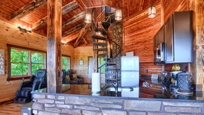 Interior de cabana com lareira de pedra, vigas de madeira no teto, parede de pedra, cozinha e área de estar com cadeiras.