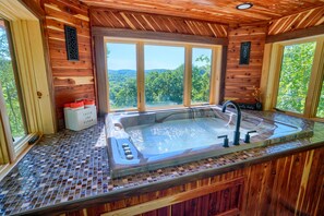 A wooden cabin with a glass-enclosed hot tub, large windows, and a view of the forest.