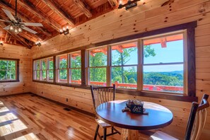 Une cabane en bois dotée de grandes fenêtres, d’un ventilateur de plafond et d’une table à manger sur laquelle se trouve un panier de fruits.