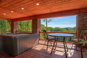 A wooden deck with a hot tub, two chairs, and a table overlooking a forested area.