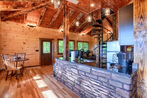 A rustic cabin interior with wooden walls, a stone counter, a dining area, and a staircase leading to a loft.
