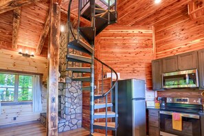 Ein Blockhaus-Interieur mit Wendeltreppe, Steinwand, Holzböden, einer Küche mit modernen Geräten und einem Fenster mit Vorhängen.