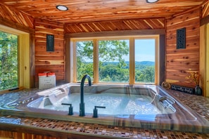 Eine Holzdiele mit einem gläsernen Whirlpool, großen Fenstern und Blick auf den Wald.