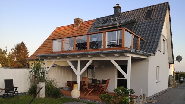Une maison à deux étages avec un toit en pente, un balcon doté de rampes en verre et un patio aménagé avec du mobilier d’extérieur.