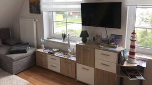Un salon confortable avec un téléviseur, une bibliothèque, une fenêtre avec des stores, une lampe et un phare décoratif.