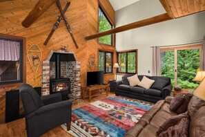 Una sala de estar acogedora con chimenea, un sofá, un televisor y un amplio ventanal con vistas al follaje.