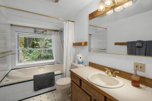 Une salle de bain moderne avec une baignoire, des toilettes et un lavabo. Il y a une fenêtre donnant sur des arbres, un miroir avec éclairage intégré et un porte-serviettes.
