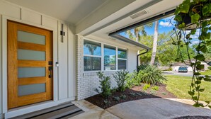 A modern house with a wooden front door, white brick walls, and a covered porch with plants. - Lux Pool Home Close to Mayo & Jacksonville Beach! (Jacksonville)