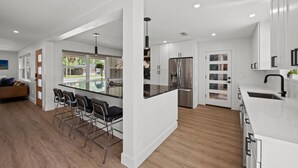 A modern kitchen with white cabinetry, a central island with bar stools, stainless steel appliances, and a large window providing natural light. - Lux Pool Home Close to Mayo & Jacksonville Beach! (Jacksonville)