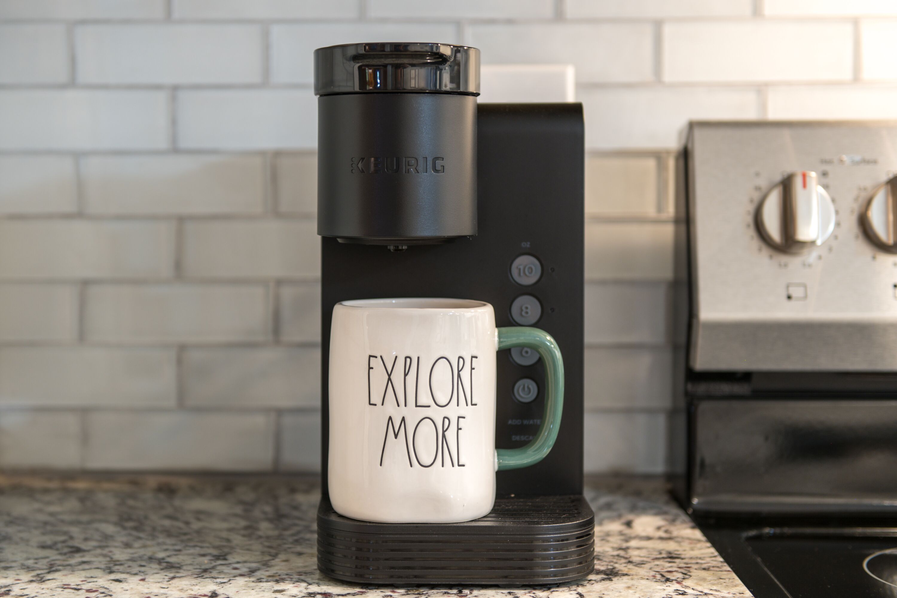 A coffee maker with a mug labeled "EXPLORE MORE" on a kitchen counter.