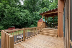 A wooden deck with a covered seating area, surrounded by lush greenery. - Private Cabin + Screened Porch + Firepit + Deck (Sevierville)