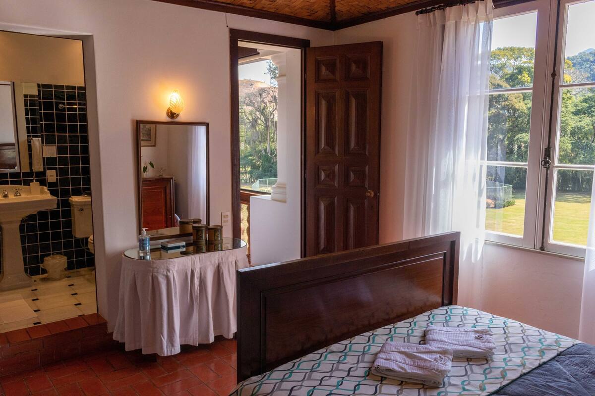 A bedroom with a bed, a bedside table, a mirror, a door, and a window with a view of greenery.