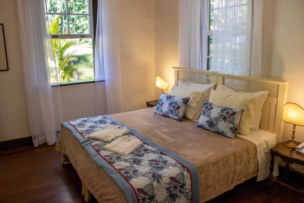 A neatly made bed with a floral bedspread, two bedside lamps, and a view of greenery through the window.