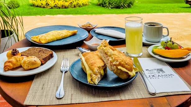 Una mesa con alimentos para el desayuno, como cruasanes, un sándwich y un vaso de jugo, con un exuberante paisaje verde y un cielo despejado de fondo.