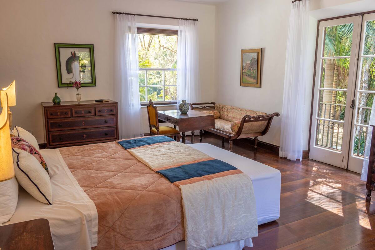 A bedroom with a large bed, a wooden dresser, a chair, a small table, and a window with curtains.