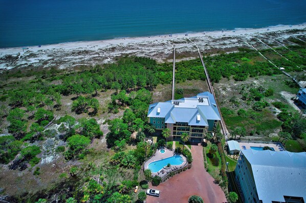 An aerial view of a hotel with a swimming pool, surrounded by lush greenery and a beach.