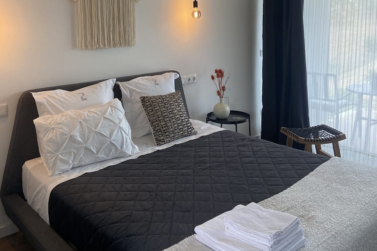 A neatly made bed with a black quilt, white pillows, and a folded towel. A macrame wall hanging is above the bed, and a small table with a vase of flowers is beside it.