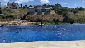A swimming pool with a person in the water, surrounded by a hilly landscape and residential buildings. - Zambujal Suites (Sesimbra)