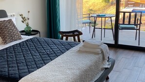 A modern bedroom with a large bed, a small table with a vase, and a view of a deck and outdoor area. - Zambujal Suites (Sesimbra)