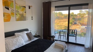 A bedroom with a bed, a bedside table, and a view of a balcony with a table and chairs. - Zambujal Suites (Sesimbra)