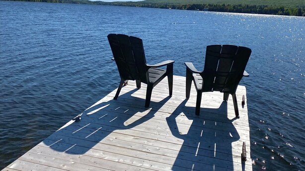 Une jetée en bois avec deux chaises, donnant sur un lac paisible, entourée de collines verdoyantes sous un ciel dégagé.