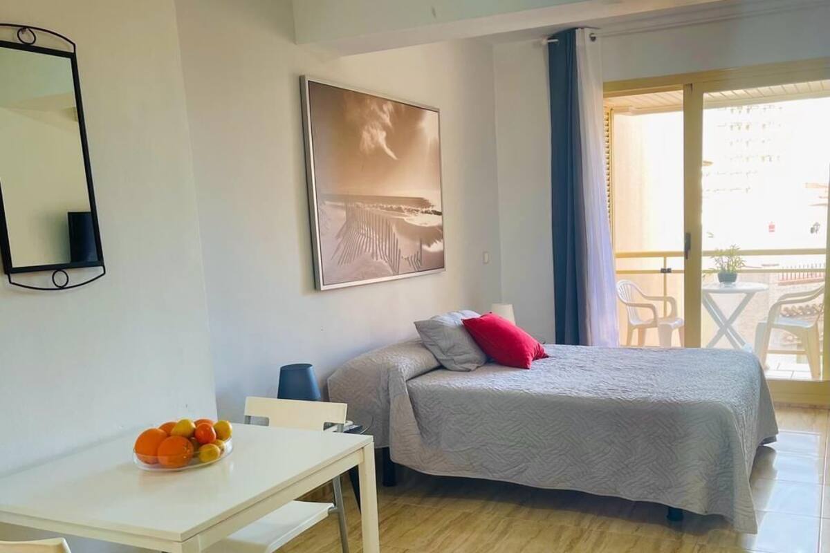 Habitación de hotel con cama, escritorio, silla, una pequeña mesa con fruta y balcón con sillas.