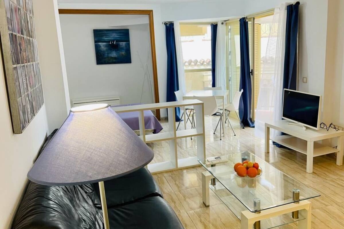 Una sala de estar moderna con una mesa de centro de vidrio, un sofá de cuero negro y un televisor sobre un mueble bajo.