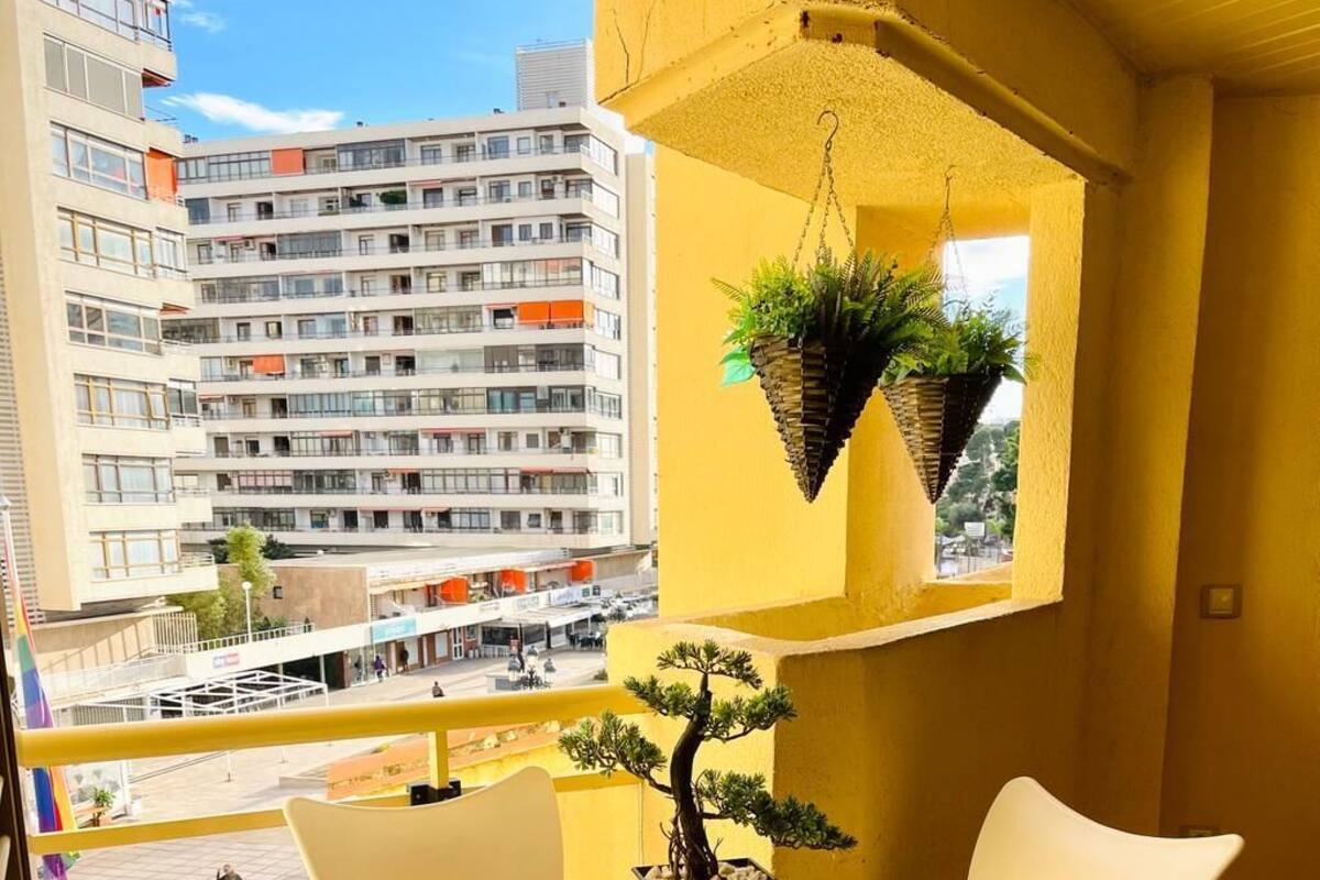 Un balcón con una mesa y sillas blancas, un árbol bonsái y macetas colgantes con follaje.