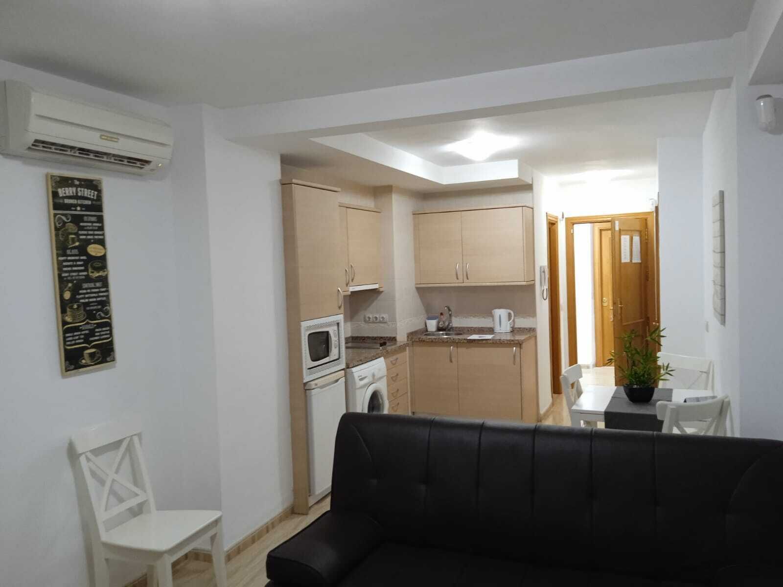 A compact living space with a kitchenette, a black leather sofa, a white chair, and a small dining table.