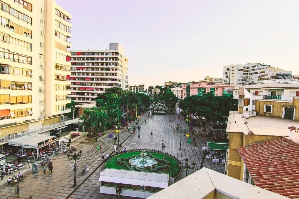 Uma praça com uma fonte, rodeada por edifícios com varandas e lojas.