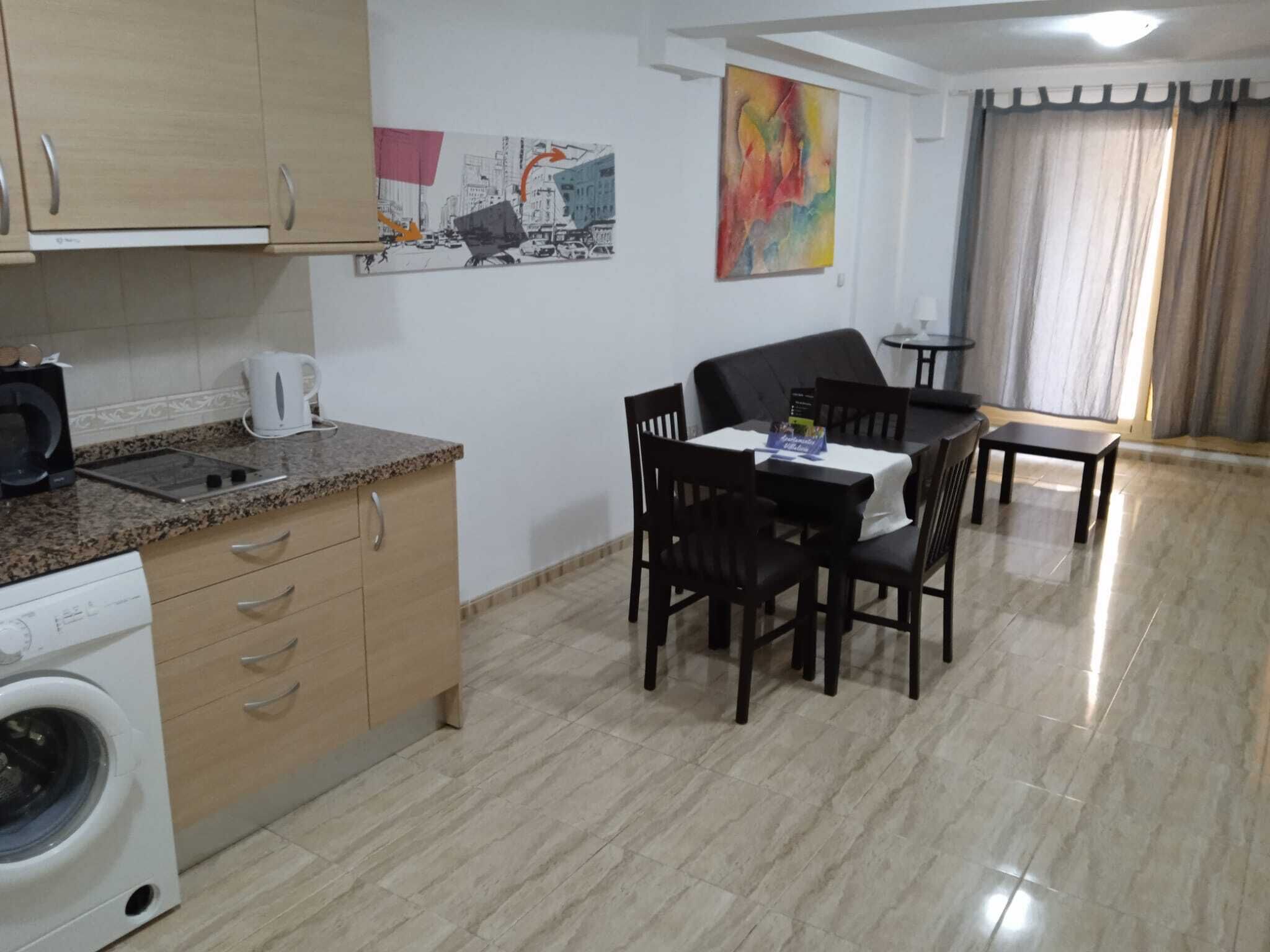 A compact kitchen with a washing machine, a microwave, and a coffee maker. There is a dining area with a table and chairs, and a colorful abstract painting on the wall.