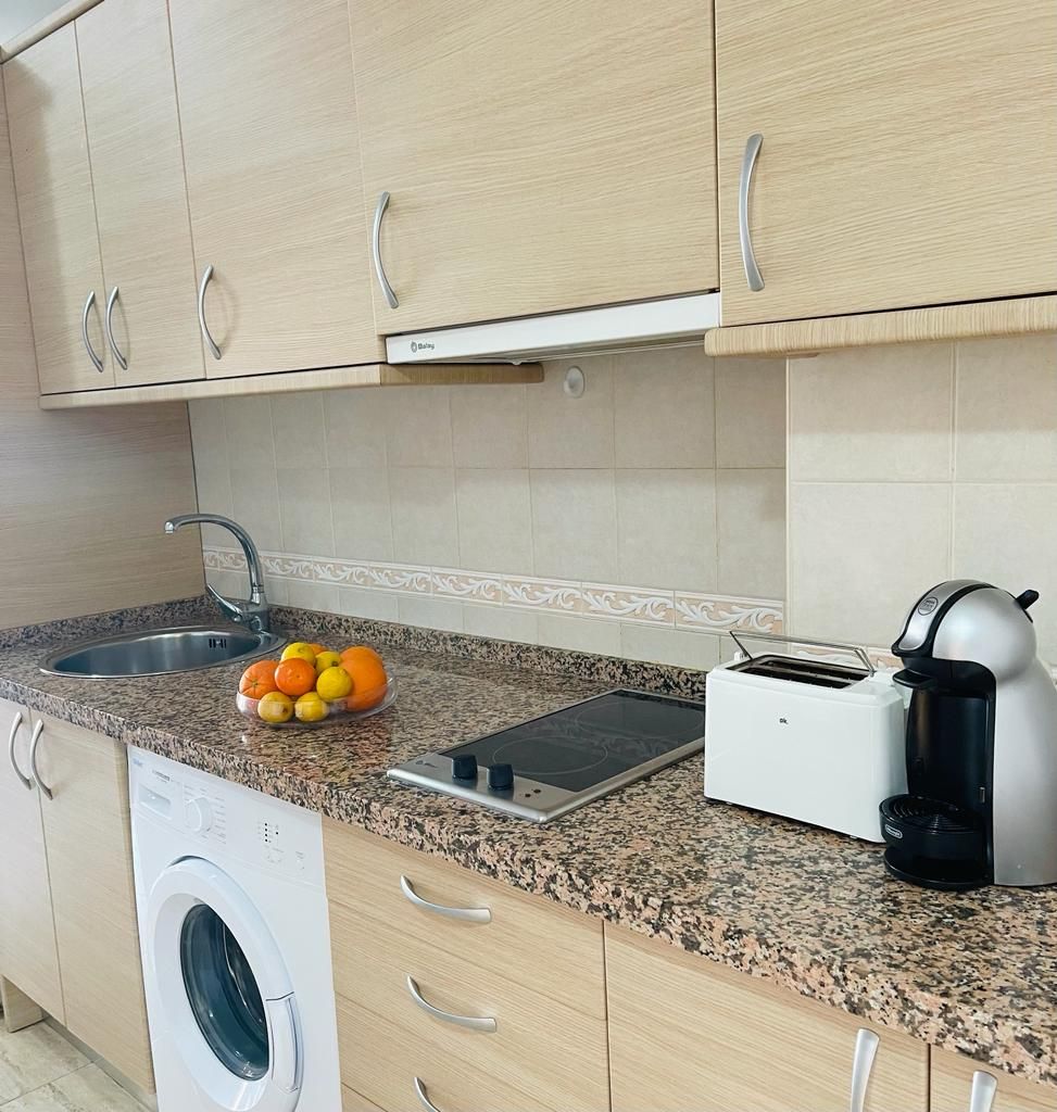 Une cuisine équipée d’un lave-linge, d’un grille-pain et d’un panier à fruits posé sur le plan de travail.