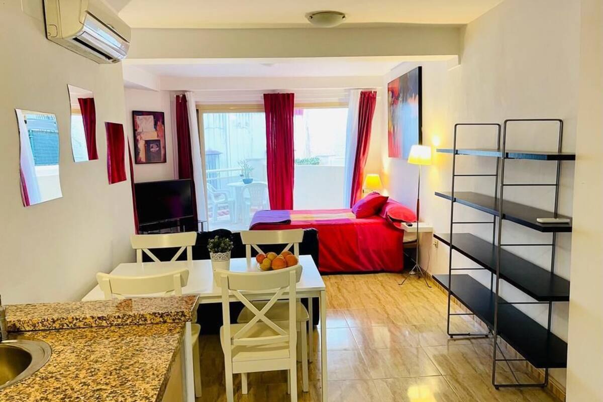 Un espacio habitable compacto con kitchenette, zona de comedor, cama con ropa de cama roja y televisor.