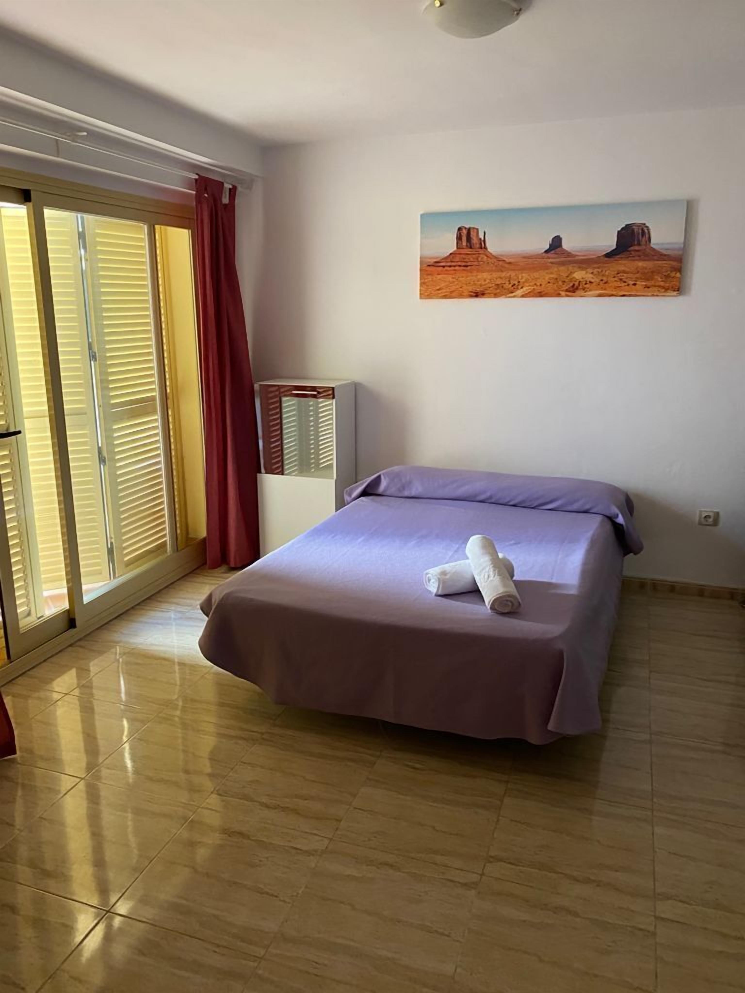 A room with a bed, a painting of a desert landscape, a heater, and a window with blinds.