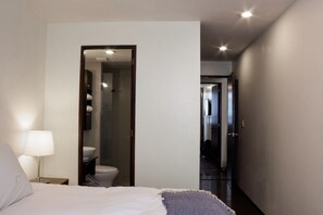 A hotel room with a bed, a nightstand with a lamp, a bathroom visible through a doorway, and a hallway leading to another room.