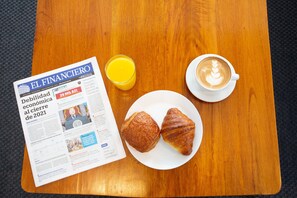 A newspaper, a glass of orange juice, a cup of coffee, and two croissants on a wooden table.