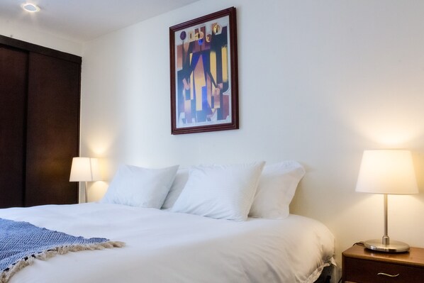 A neatly made bed with white linens and a blue throw blanket, a bedside lamp, and a framed abstract painting on the wall.