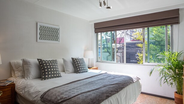 A bedroom with a large bed, a window with a view of the outside, a wall-mounted light fixture, and a framed artwork. - Mai Calli Home Rentals (Mexico City)