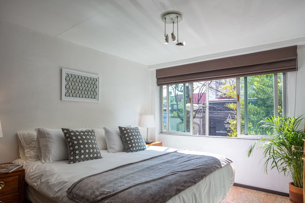 A bedroom with a large bed, a window with a view of the outside, a wall-mounted light fixture, and a framed artwork.