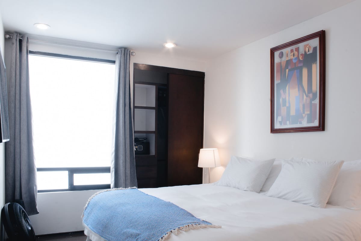 A hotel room with a bed, a bedside lamp, a window with curtains, and a painting on the wall.