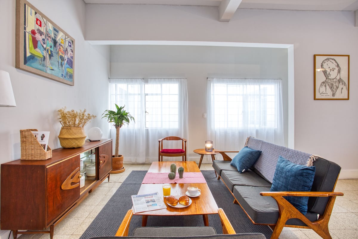 A modern living room with a sofa, a wooden table with breakfast items, a cabinet, and a large window.