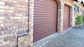 A brick building with a closed brown garage door and a small green bush near the entrance. - Spacious holiday home in cozy complex. Walking distance from the nature reserve. (KZN)