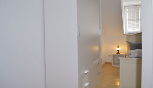 A narrow hallway with a bed and nightstand in the distance, a window with blinds, and a wall-mounted light fixture.
