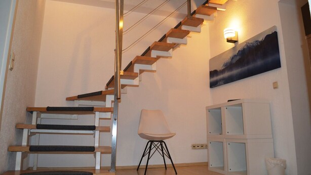 A staircase with wooden steps and a metal railing, a white chair, a cabinet with glass doors, and a wall-mounted light fixture. - City Center Stays /// AK4-L, duplex apartment (München)
