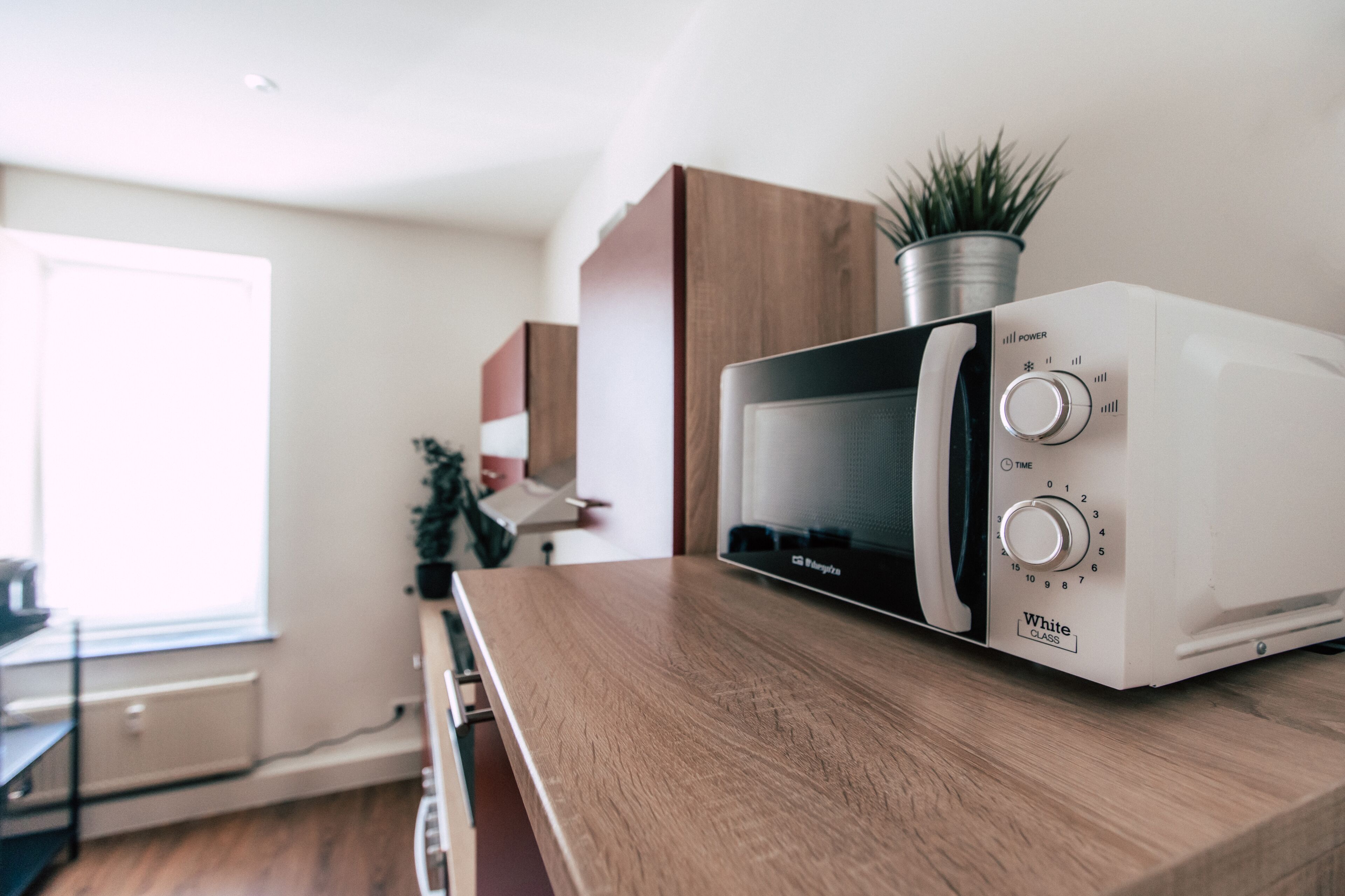 A microwave on a wooden countertop in a kitchen with a window and cabinets in the background.