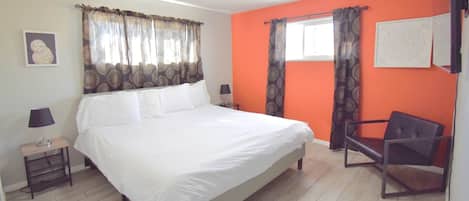 A bedroom with a large bed, a chair, a nightstand, a lamp, and a window with curtains.