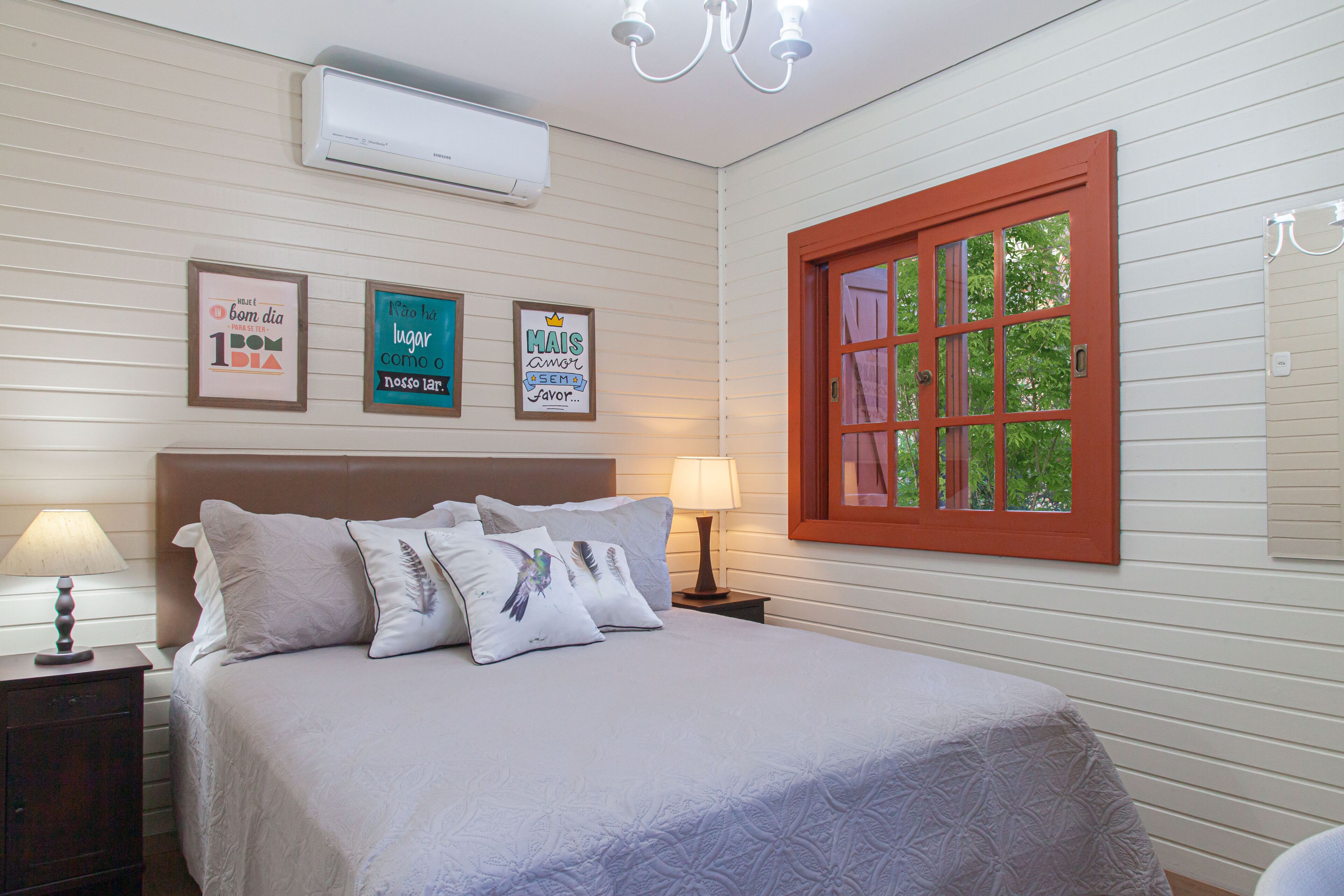 A bedroom with a bed, nightstands, wall-mounted air conditioning, framed artwork, and a window with a red frame.