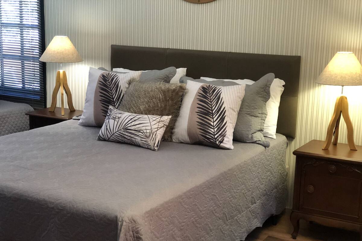 Un lit bien fait, recouvert d’une couette grise, agrémenté de plusieurs oreillers ornés de motifs de feuilles, et deux tables de chevet avec des lampes.