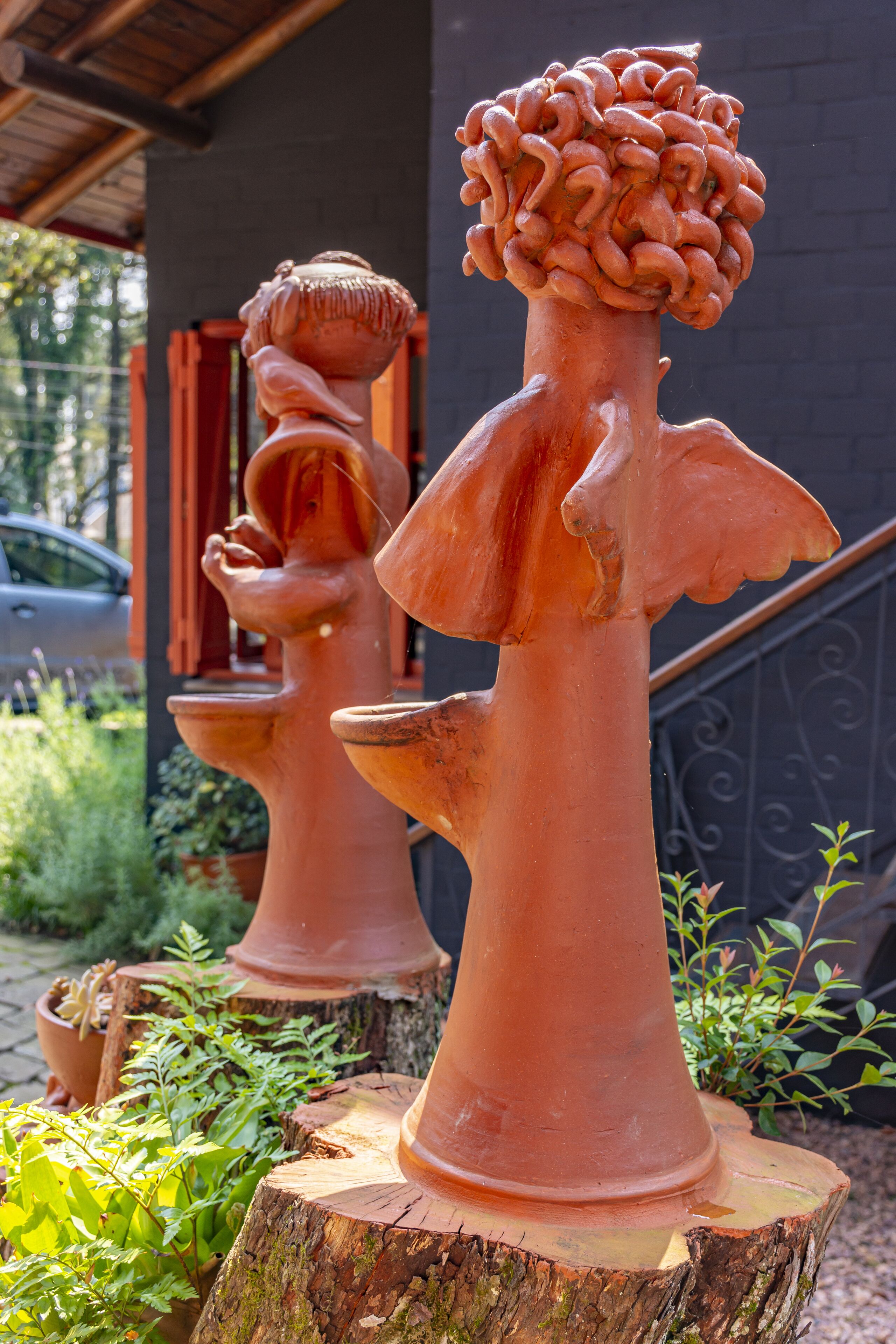 Esculturas de terracota de ángeles alados, colocadas sobre tocones de árboles en un jardín.