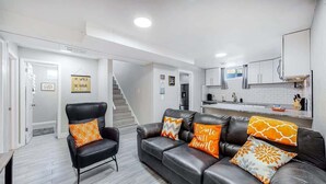 Una sala de estar moderna con un sofá y sillón de cuero negro, una escalera y una zona de cocina con armarios blancos y un fregadero de acero inoxidable.